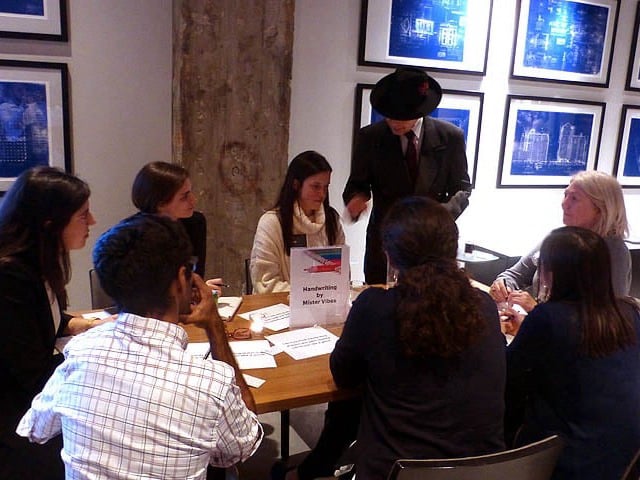 Mister Vibe analyzing the handwriting of a group seated at a table.